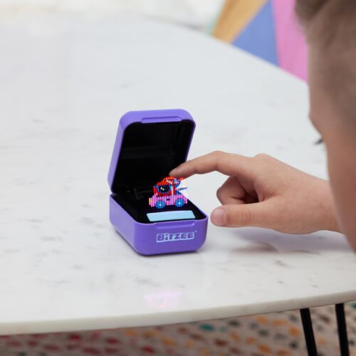 Junge interagiert konzentriert mit einem geöffneten Bitzee-Spiel, einem handlichen Lernspielzeug mit Pixelgrafik-Display auf einem weißen Tisch.